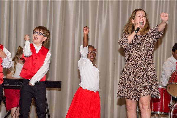 A group of children and an adult woman perform on stage. The woman, wearing a leopard print dress, sings into a microphone with one arm raised. A young girl in a white blouse and red skirt and a boy in a red vest and glasses enthusiastically raise their fists in the air. A keyboard and drum set are 